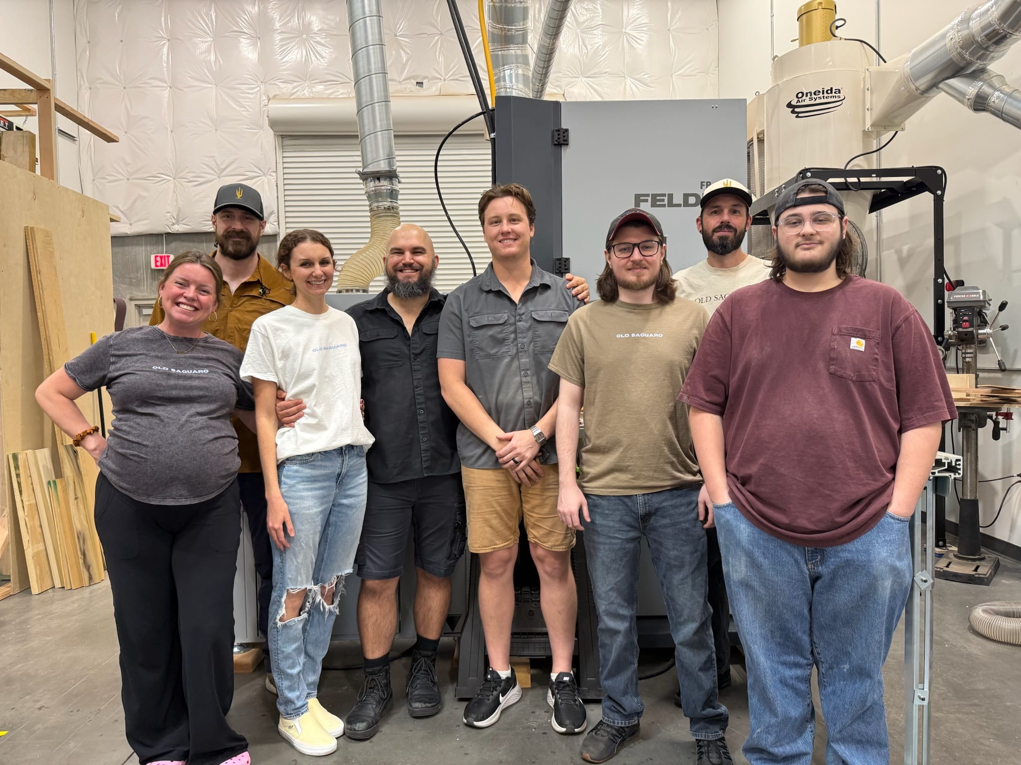 Eight people stand smiling in a workshop in front of a gray Felder machine and an Oneida dust collector, with wood stock leaning at left.
