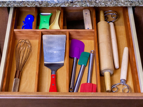 An open wooden kitchen drawer organized with colorful kitchen utensils, including a metal whisk, spatula, purple and green silicone spatulas, a wooden rolling pin, and other baking tools.